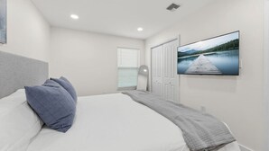 Ein Schlafzimmer mit Bett, Kissen, Fernseher, Kleiderschrank und einem Fenster mit Jalousien.