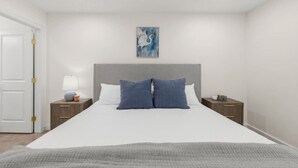 Un dormitorio con cama, dos mesitas de noche, una lámpara, un cuadro y una puerta.