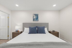A bedroom with a bed, two bedside tables, a nightstand, a lamp, a painting, and a door.