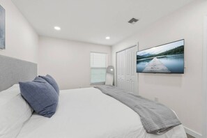 Una camera da letto con un letto, cuscini, una TV, una finestra con persiane e una porta.
