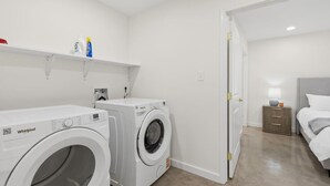 Una lavandería con dos lavadoras Whirlpool, un estante con productos de limpieza y un dormitorio con cama y mesita de noche.