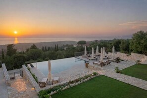 Zona junto a la piscina con sillones y sombrillas, terraza de piedra y vista al mar al atardecer.