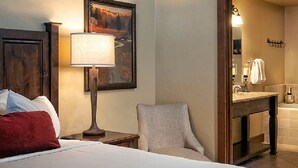 Habitación de hotel con cama, una silla, lámpara, un cuadro, y un baño con lavamanos y espejo.