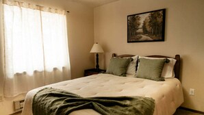 A bedroom with a bed, a nightstand, a lamp, a window with curtains, and a painting on the wall. - 3 bedroom Townhome in West Yellowstone, perfect for families!!! (West Yellowstone)