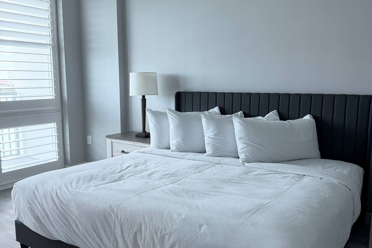 A modern bedroom with a large bed, a bedside table with a lamp, and a window with blinds.
