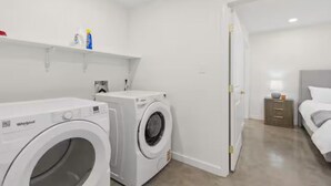 Une buanderie équipée de deux lave-linge Whirlpool, d’une petite étagère contenant des produits de nettoyage et d’une porte donnant sur une chambre avec un lit et une table de chevet.