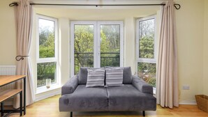 Una habitación con un sofá gris, una mesa de madera y ventanas grandes con cortinas.