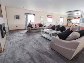A spacious living room with a grey carpet, two sofas with black and red cushions, a coffee table, and a kitchen in the background.