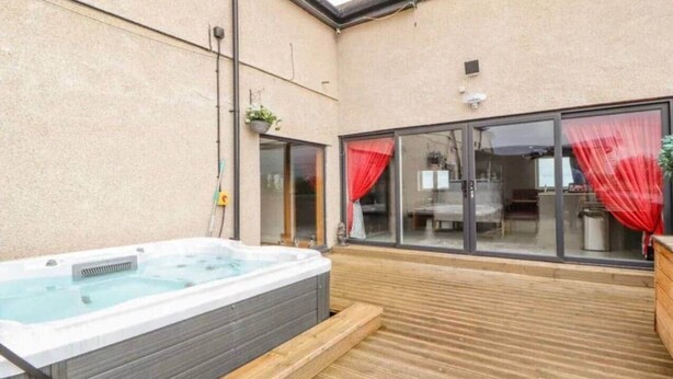 A hot tub in a covered outdoor area with a wooden deck, a sliding glass door, and a window with red curtains. - Penthouse apartment in the heart of towyn (Towyn)