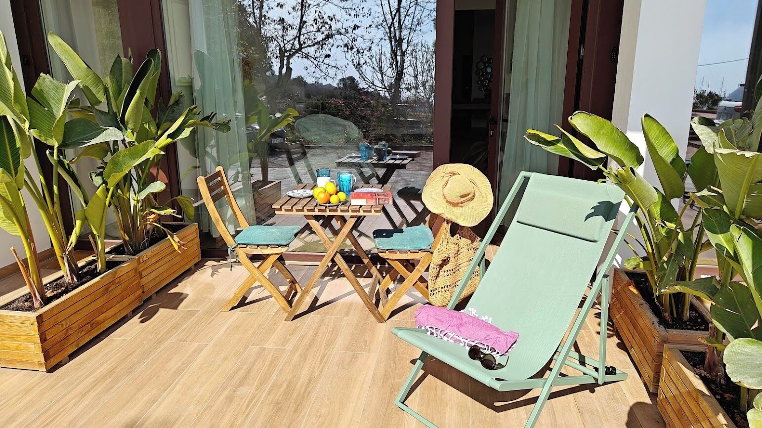 An outdoor seating area with wooden tables and chairs, a sun lounger, and potted plants.