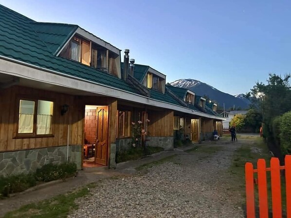 A row of wooden cabins with stone foundations, a gravel path, and a mountain in the background. - Apart Hotel Queitao (Puerto Aysen)