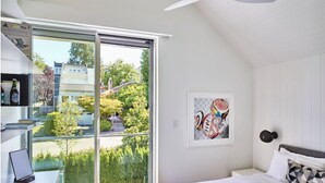 Een slaapkamer met een bed, een bureau met een laptop, een stoel, een raam met uitzicht op groen en gebouwen, en een plafondventilator.