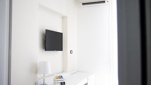 Une pièce blanche avec un téléviseur à écran plat fixé au mur, un bureau blanc avec une lampe et une petite pile de livres.