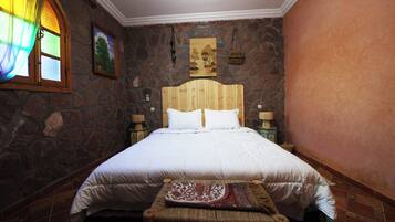 A bedroom with a stone wall, a wooden headboard, a bed with white bedding, a nightstand with a lamp, and a small bench with a cushion.