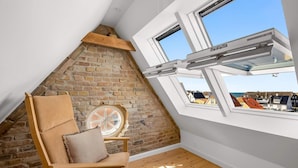 A room with a sloped ceiling, a brick wall, a wooden chair, and a window overlooking a town. - 4 Star Holiday Home in Lokken (Lokken)