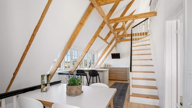 A modern loft with a wooden beam ceiling, a dining area with white chairs, a kitchen with wooden cabinets, and a staircase. - 4 Star Holiday Home in Lokken (Lokken)