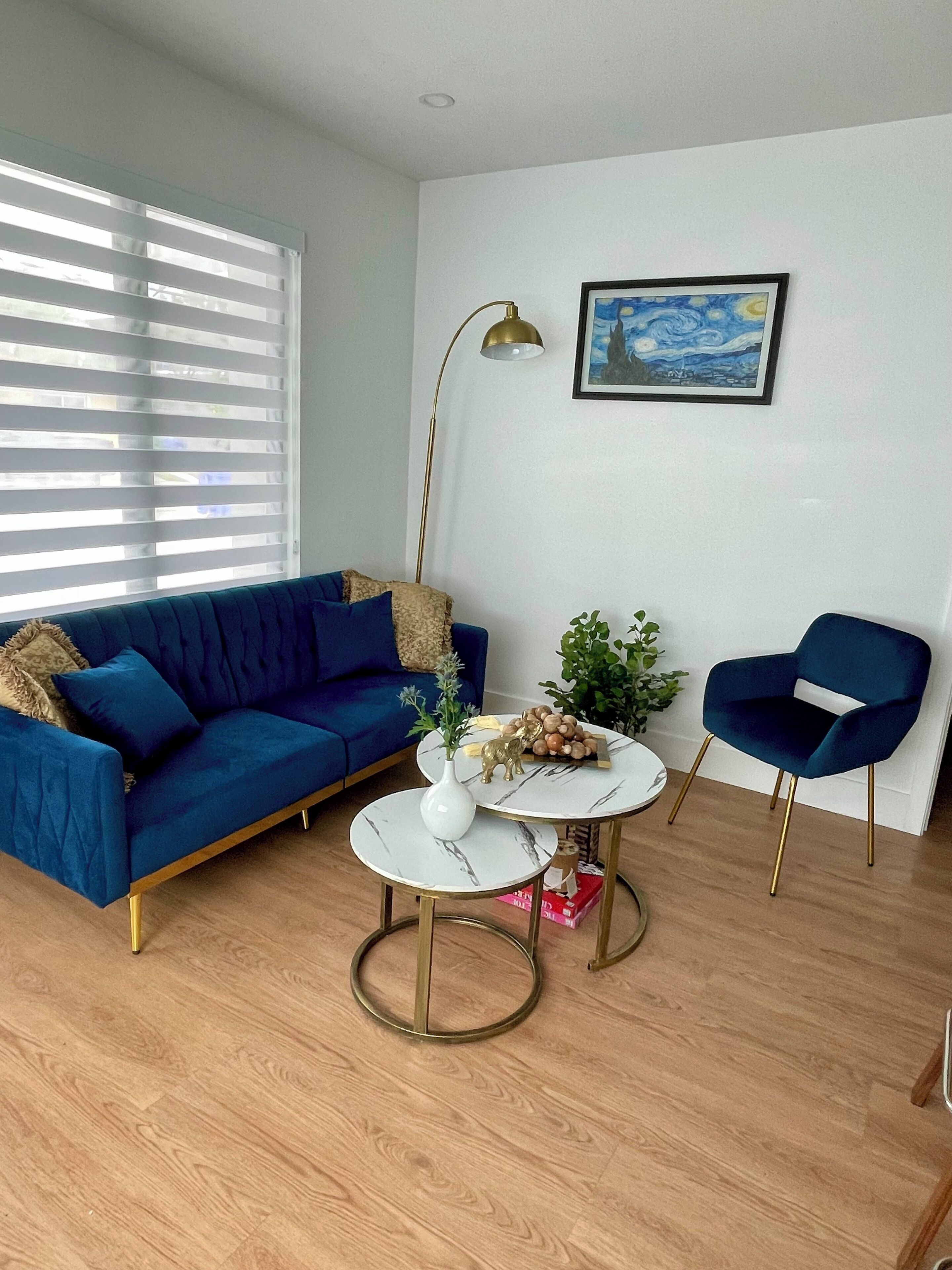 Ein Wohnzimmer mit einem blauen Sofa, einem runden Couchtisch, einem blauen Sessel, einer Stehlampe und einem Gemälde an der Wand.