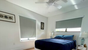 Un dormitorio con cama, ventilador de techo, ventana y mesita de noche.