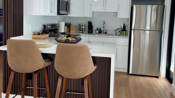 Una cocina moderna con armarios blancos, un refrigerador de acero inoxidable, dos taburetes de bar e iluminación colgante.