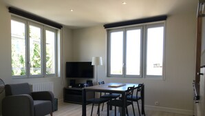 A modern living room with a flat-screen TV, a dining table with chairs, a sofa, and large windows.