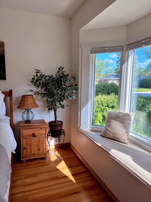 Et soveværelse med et trænatbord, en lampe, en potteplante, et vindue med udsigt til grøn vegetation og en polstret bænk.