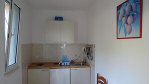A compact kitchen with white cabinets, a sink, and a small refrigerator. There is a window on the left and a chair at the table. - Villa Azure Saplunara - One bedroom apartment (Saplunara)