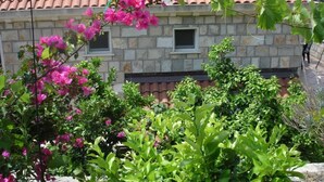 A stone building with a tiled roof, two small windows, and lush greenery including pink flowers. - Villa Azure Saplunara - One bedroom apartment (Saplunara)