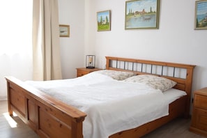 Una cama de madera con una colcha blanca y dos almohadas, una mesita de noche con una lámpara, y una pared con tres cuadros enmarcados.