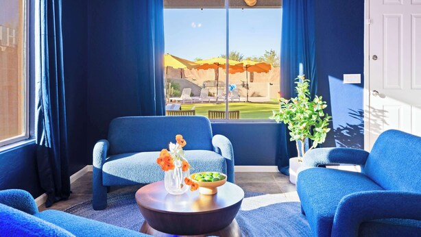 Una habitación con dos sillones azules, una mesa redonda con un jarrón y un cuenco, una planta en maceta, y vistas a un espacio exterior con sombrillas.