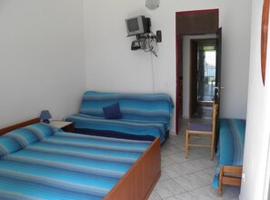 Ein Einzelbett mit gestreifter blau-weißer Bettdecke, ein kleiner Nachttisch mit Lampe und ein an der Wand befestigtes Fernsehgerät.