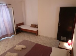 Ein schlichter, kleiner Raum mit einem Bett, einer Schlafcouch, einer Tür und einem Fenster mit Vorhängen.