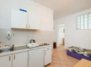 Eine kleine Küchenzeile mit Spüle, Wasserkocher und Mikrowelle. Ein Bett mit bunter Decke und ein Fenster mit Blick auf Bäume.