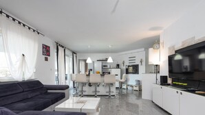 Una sala de estar moderna con sofá, mesa de centro y área de cocina con televisor, taburetes de bar y mesa de comedor.