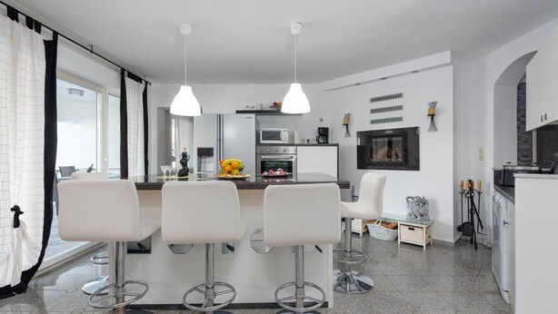 Una cocina moderna con una isla central, taburetes de bar, iluminación colgante y un comedor con sillas.