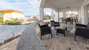 Un patio al aire libre cubierto con suelo de piedra, muebles de mimbre negro y vistas al mar y edificios.