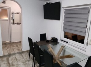A dining area with a glass table, black chairs, a black speaker, and a window with blinds. - Two Bedroom Apartment with Terrace (Vir)