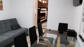 Un soggiorno moderno con un divano grigio, un tavolo da pranzo con piano in vetro, sedie nere e una TV a parete.
