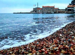 A coastal scene with a pebbled beach, a building with a red roof, and a body of water. - Villa Teodora Mare - Studio with Sea View (App 2) (Petrovac na Moru)
