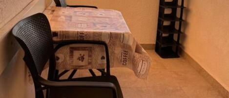 Un balcon avec une table recouverte de nappes aux motifs de café, deux chaises et une petite étagère.