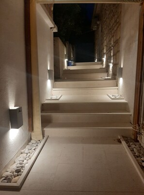 A staircase leading to a darkened upper level, with lights along the steps and walls, and a stone wall on one side. - Apartments Trojanović - Three Bedroom Apartment with Sea View and Balcony (Orebic)