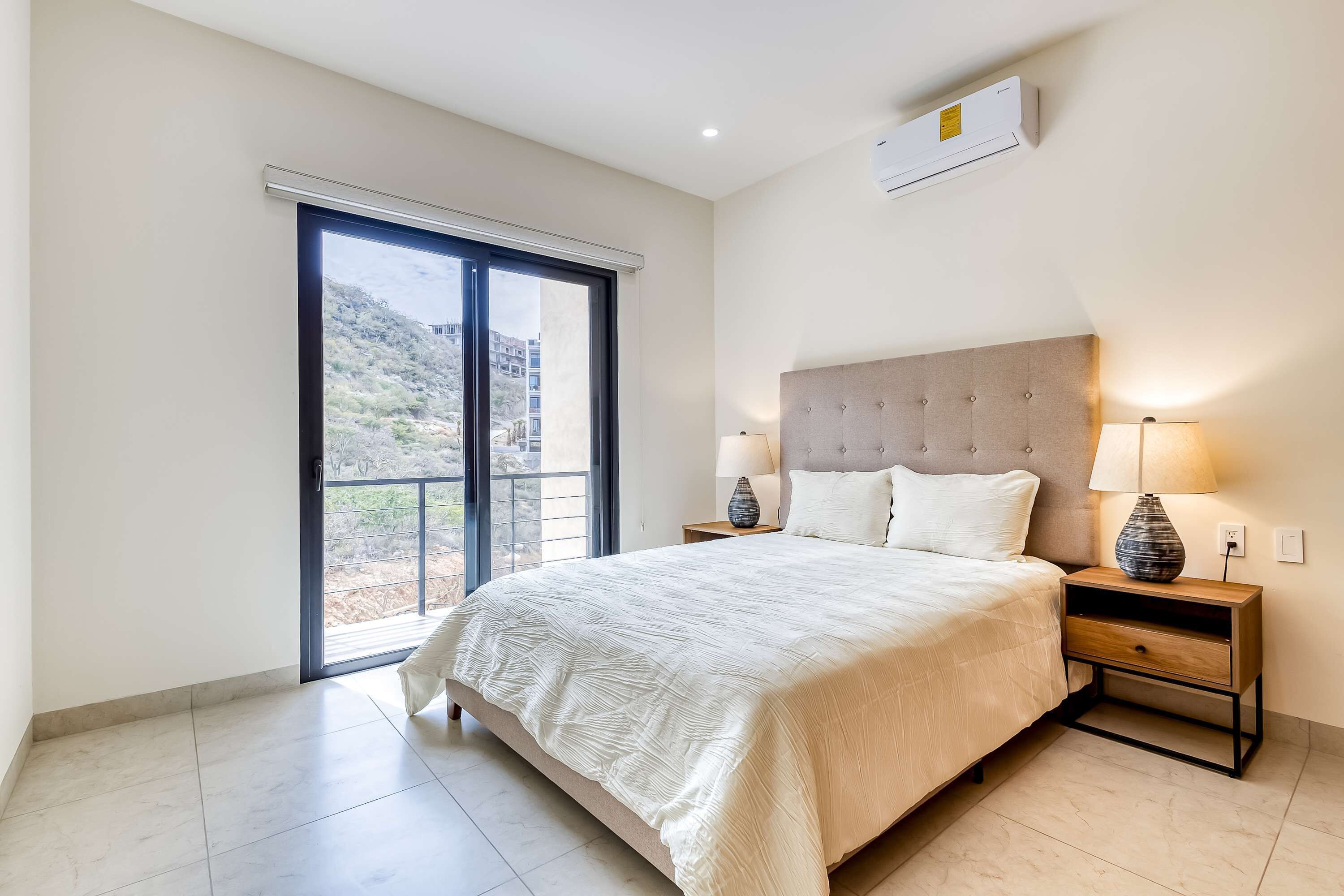 Un dormitorio con cama, mesitas de noche, lámpara, aire acondicionado y puerta corrediza a un balcón.