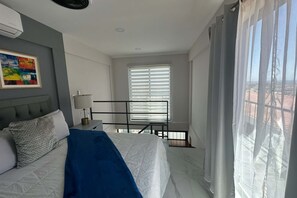 Een slaapkamer met een bed, nachtkastje, lamp, raam met zonwering en uitzicht op buiten.