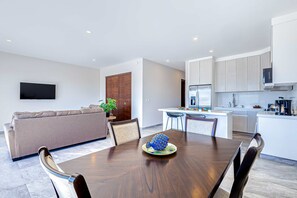 A modern living room with a sofa, dining table, and chairs, a kitchen area with white cabinets, and a flat-screen TV mounted on the wall. - Stunning Oceanfront Luxury Apartment with spacious balcony– Perfect for Families (Cabo San Lucas)