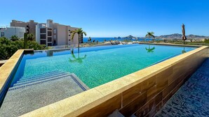 Ein Schwimmbecken mit klarem Wasser, einer gefliesten Terrasse und Blick auf das Meer und Gebäude im Hintergrund.