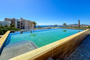 A swimming pool with clear water, a tiled deck, and a view of the sea and buildings in the background. - Stunning Oceanfront Luxury Apartment with spacious balcony– Perfect for Families (Cabo San Lucas)