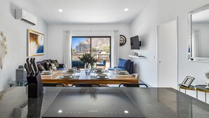 Un espace de repas moderne avec une table dressée pour quatre personnes, un îlot de cuisine doté d’une plaque de cuisson à induction, une table à manger avec des chaises, un canapé et une vue sur un paysage montagneux par la fenêtre.