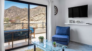 Un salon moderne avec une table basse en verre, un canapé bleu, un téléviseur fixé au mur et un balcon offrant une vue sur un paysage montagneux.