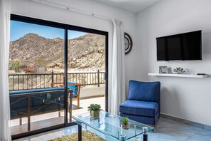 Ein modernes Wohnzimmer mit einem Glastisch, einem blauen Sofa, einem an der Wand montierten Fernseher und einem Balkon mit Blick auf eine gebirgige Landschaft.