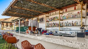 Een bar met een counter, barstoelen, flessen op planken en uitzicht op een heuvelachtig landschap.