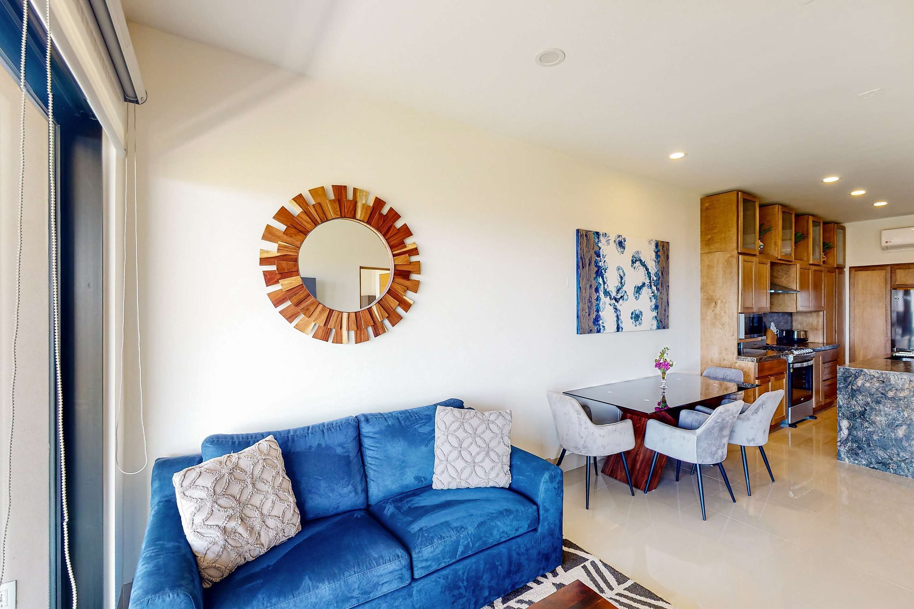 A modern living room with a blue sofa, a dining table with chairs, a kitchen area, and a large mirror on the wall.
